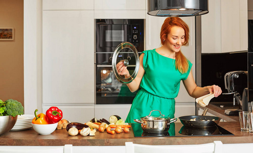 红头发站在厨房的桌子，与五颜六色的蔬菜，帽壶和钳在高科技现代阳光厨房附近的漂亮微笑殷勤女人