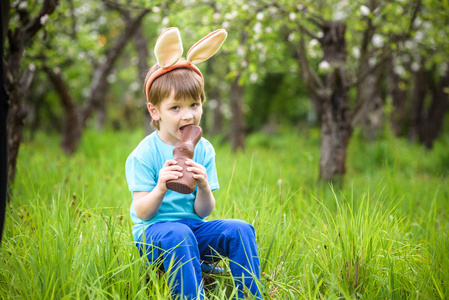 快乐小蹒跚学步的男孩吃巧克力和头戴复活节兔子耳朵，在盛开的花园里坐在暖和的晴天。庆祝传统节日