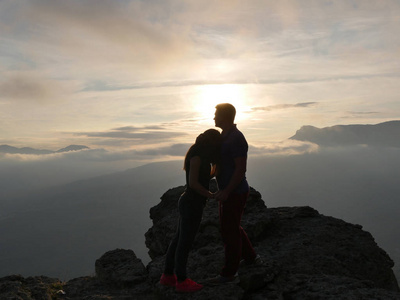 年轻夫妇站在一座山和看彼此在美丽的日落背景的剪影。男孩和女孩的爱