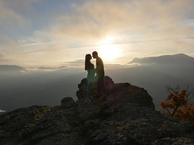 年轻夫妇站在一座山和看彼此在美丽的日落背景的剪影。男孩和女孩的爱