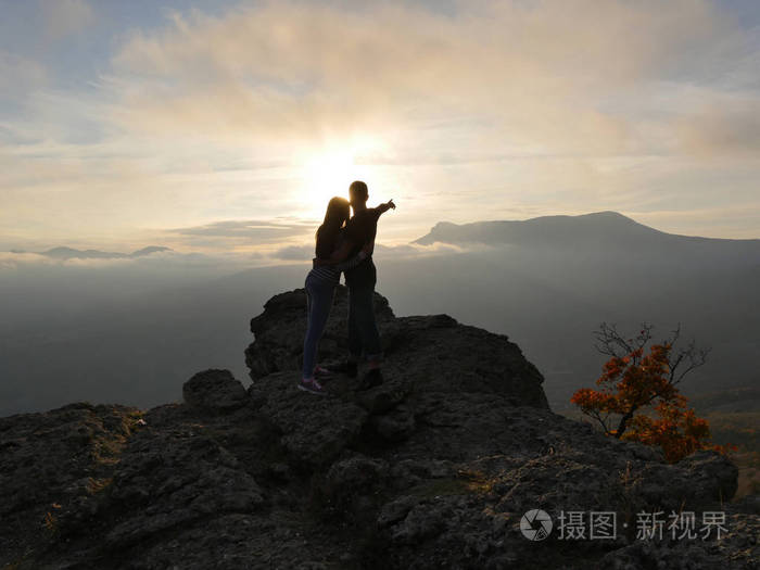 年轻夫妇站在一座山和看彼此在美丽的日落背景的剪影男孩和女孩的爱