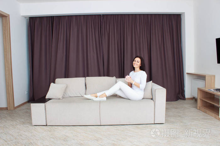美丽的年轻女子坐在沙发上在家里与杯茶的肖像