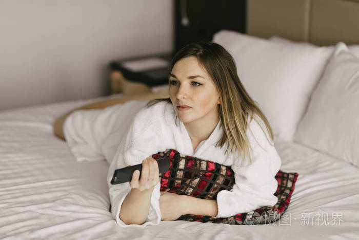 美丽的女人躺在床上和在酒店房间看电视