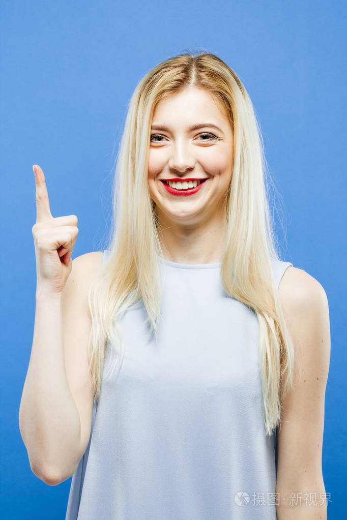 笑着的女孩是显示了她的手指,看着相机在工作室中的蓝色背景上