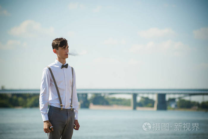 年轻的瘦男人,优雅时髦穿着白衬衫,灰色长裤吊带及领结