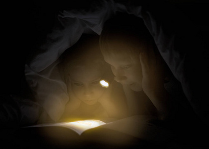 姐姐和弟弟正在阅读一本书下毛毯，手电筒。年轻漂亮的男孩和可爱的女孩在儿童房开心