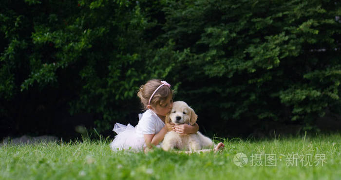 一个小女孩打扮成小舞蹈家，亲吻她的小小朋友小狗的狗黄金猎犬坐在草坪和幸福的概念友谊 狗和人类之间的友谊。连接