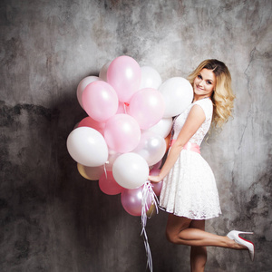 迷人的年轻金发女郎穿着白色与粉红色的腰带，抱着一大捆的气球。在灰色带纹理的背景上
