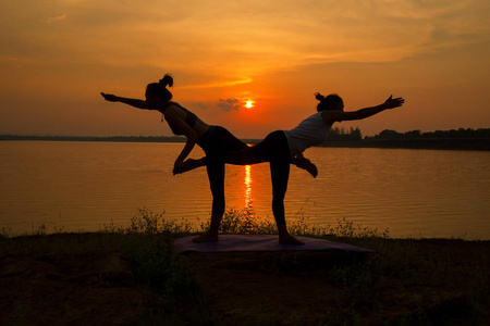 练习瑜伽体式上日落河流的瑜伽剪影两个女人