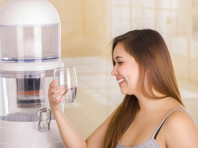 美丽微笑的女人，拿着一杯水，与水净化器厨房背景上过滤系统