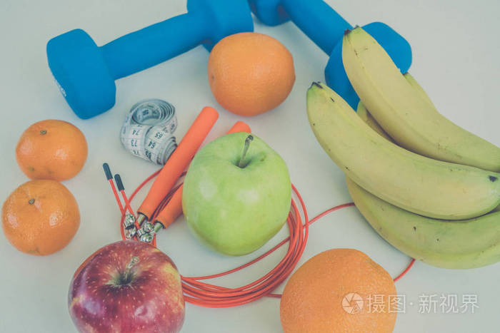 健康饮食的概念。哑铃 绳 香蕉 橘子 赌 橘子 红苹果 青苹果 橙汁白色的桌子上。健康的生活方式。运动。健身