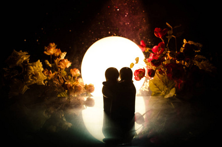 两个娃娃拥抱用鲜花和月亮装饰光背景与烟的桌子上。爱的概念。问候或礼品卡设计理念。老式的口气。拥抱夫妇的剪影