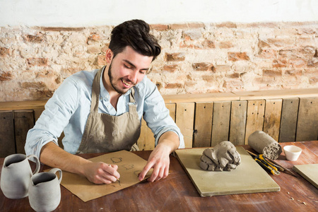 波特，车间，工匠工具，陶瓷艺术概念微笑男性年轻人对未来产品的设计工作与工具 粘土 成品的罐坐在桌子后面的陶艺家
