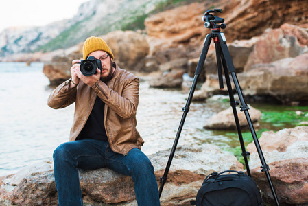 年轻时尚的摄影师，胡子，戴着黄色的帽子和眼镜，坐在岩石上，为你拍照。 他旁边的三脚架和背包。 地中海