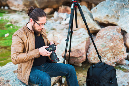 年轻时尚的摄影师戴着眼镜，胡子坐在岩石上，看着他在相机上拍的照片。 他旁边的三脚架和背包。