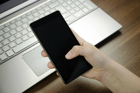 用手指拿黑色的智能手机躺在木垫模糊的银笔记本电脑键盘上的一个女人的手的特写视图