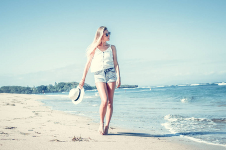 年轻的女人与一个热带的巴厘岛在阳光明媚的日子走在白色的沙滩上的白帽子。海洋成本