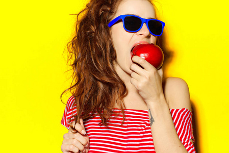 美丽年轻的女孩在蓝色太阳镜和红色条纹 t 恤在黄色背景上吃苹果又开心又笑