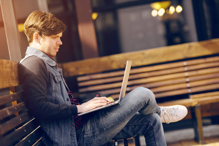 年轻的家伙坐在板凳上大街上，并使用一台笔记本电脑。生活方式和技术的概念