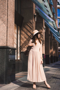 苗条的年轻女孩博主的肖像在市中心美丽的黑妞杜塞尔多夫的柔和的裙子和一位女士的帽子戴着墨镜，走路的姿势和日落微笑