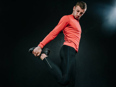 底部视图的欧洲年轻人穿着红色运动服和拉伸后，在黑暗的背景中一个重锻炼他的腿。强大的英俊竞技雄性做合适。体育动机。健康的生活方式