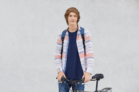年轻骑自行车的人穿着衬衫和牛仔裤，拿着背包站在他的自行车去附近的时髦发型要步行穿过城市。手牵着手在现代自行车的把手上的时髦家伙