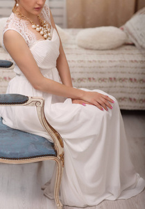 婚礼准备。年轻貌美不可识别的新娘在室内穿着白色的婚纱。坐在老式蓝色椅子上在家里在工作室里大窗口的 Luxuty 模型