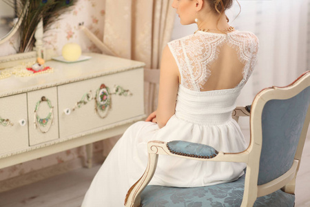 婚礼准备。在室内穿着白色婚纱的美丽年轻的新娘。坐在老式椅子上在家中工作室的豪华模型