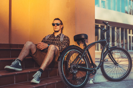 踏单车的英俊男子用背包自行车自行车附近的台阶上坐着休息旅游概念的太阳镜