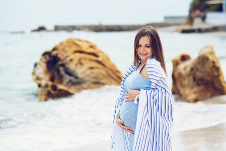 美丽的孕妇在一件紧身的衣服布满了摸她的肚子与海滩上的白色条纹格子的蓝色爱和关心的岩石和海背景