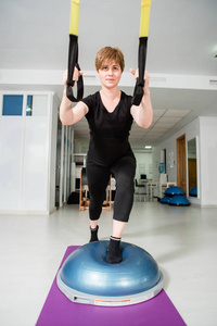 运动吸引人的年轻女子使臀部腿TRX运动站在健身房的健身球上。 背景上的其他普拉提设备。 积极健康的生活方式观念。