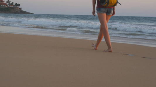 年轻女人旅行背包在日出期间沿着海滩走的腿。英尺的女游客在日落时分赤脚走在海边上。女孩的徒步旅行者旅行或夏天假期步进