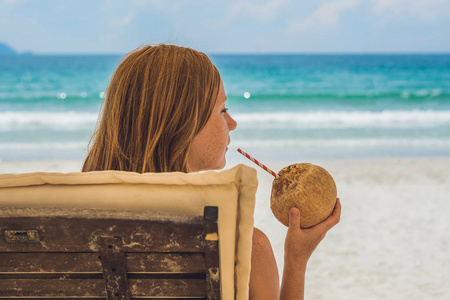 年轻女子在沙滩上的贵妃椅上喝椰子牛奶