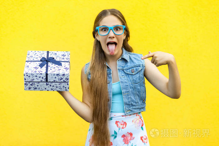 有趣的金发女孩的裙子和蓝色的眼镜，吐舌笑脸望着镜头指向手指放在礼物盒上的孤立在黄色背景。室内影棚拍摄