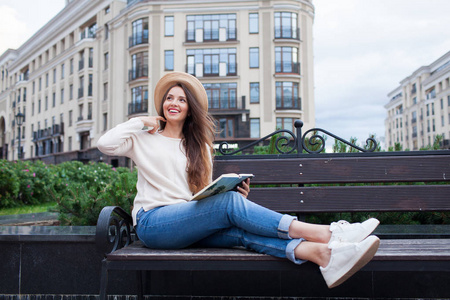 一个年轻漂亮的女人，在优雅的帽子坐在长凳上的一个新的居民区和读纸质书。她快速翻页和微笑。城市背景