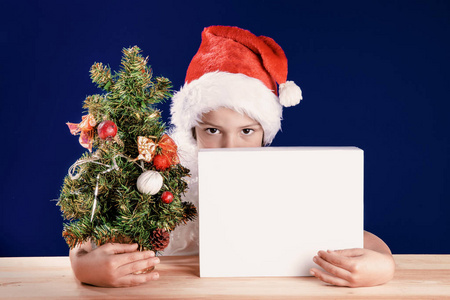 一个小男孩打扮成圣诞老人礼品包装盒外偷看。他看着镜头。在一个白色的盒子里的礼物。在蓝色的背景。特写