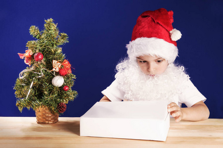 一个小男孩在圣诞老人的西装看上去带着圣诞礼物盒上。在白色的 t 恤。在一个白色的盒子里的礼物。在蓝色的背景。特写