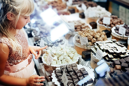 一个金发碧眼的小姑娘站在商店橱窗里着各式各样的糖果附近。孩子选择糖果