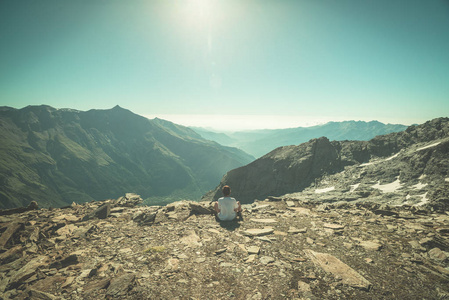 一个人坐在岩石地形上，看着色彩斑斓的朝阳在阿尔卑斯山的高处。宽视角从上面与发光的山峰在背景中。色调的图像