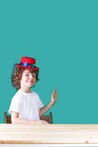 一件白衬衫和深红色帽子小丑坐在一张桌子的小卷发男孩半转身，举起手，手掌向前，着相机的镜头。特写镜头。蓝色背景