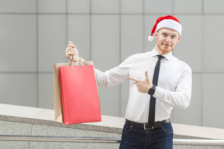 黑色星期五在冬天。幸福红头商人在圣诞老人的帽子，手上提着购物袋，指着，看着相机