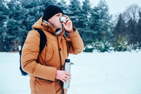 一个年轻男子在红色外套，站在雪地上，抱着一个保温瓶和一杯茶一个背包。冬季森林