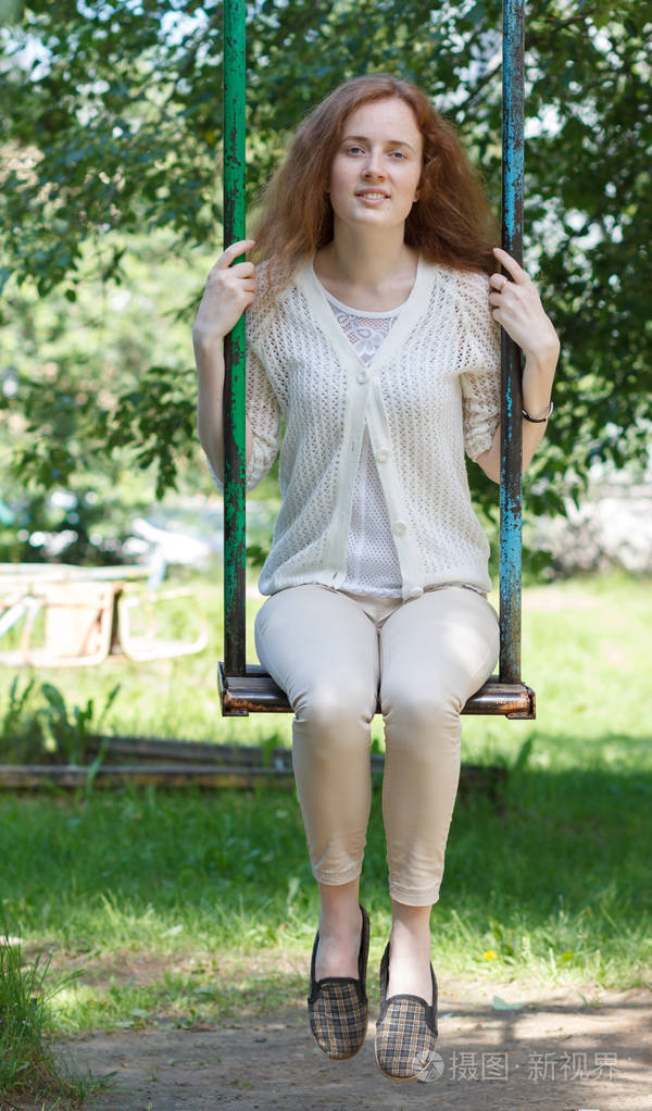 美丽年轻的红头发女人骑在一个城市公园的秋千上