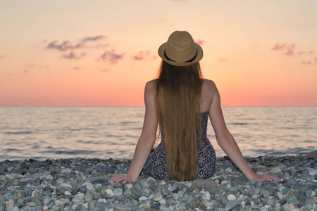 坐在海边上的这顶帽子的女孩.日落的时候.查看从背面照片