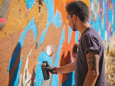 英俊有才华年轻男孩正在用气溶胶喷雾的七彩涂鸦做城市街道墙上。电影 tonedshot。创造性的艺术。侧视图