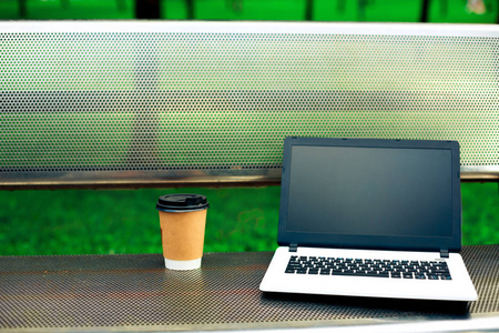 空白的黑屏和金属在自然户外公园长凳上的咖啡杯的笔记本电脑样机的形象
