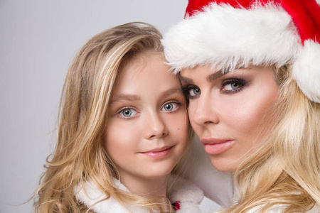 美丽的金发碧眼女模特母亲和女儿在一顶红帽子与在白色的皮毛可爱化妆打扮成圣诞老人在圣诞节的时候