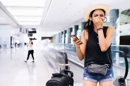 机场女人在门口等待在终端的智能手机。空中旅行概念与年轻休闲时髦女人站着随身携带的手提行李手推车。漂亮的年轻混的血女性