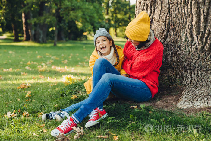 有趣的小孩和她的母亲在树附近坐在一起,走在秋天的公园,一起享受美好