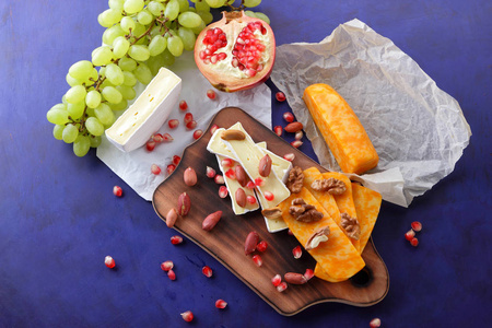 卡门培尔奶酪奶酪 橙色的硬奶酪 绿葡萄 花生 核桃 石榴种子在木板上的复古风格的深蓝色背景上
